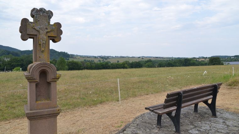 A bench and a stone cross in a rural landscape with sweeping views over fields and hills.