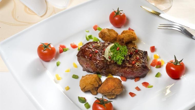 Een sappige steak op een wit bord, gegarneerd met verse tomaten en kleurrijke groenten. Daarnaast bevinden zich krokante bijgerechten.