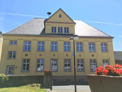 Ein gelbes Gebäude mit der Aufschrift „MUSEUM“ an der Vorderseite. Es hat ein schräges Dach und ist von grüner Landschaft und Blumen umgeben.