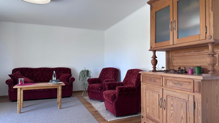 Ein gemütlicher Wohnraum mit roten Sofa- und Sesselgruppen. Ein Holzmöbelstück steht an der Wand, dazu ein kleiner Tisch und ein Teppich auf dem Boden.