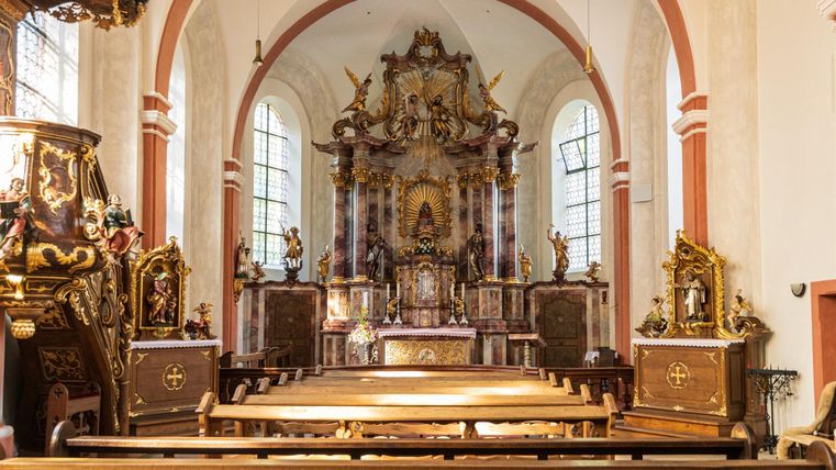 Ein schöner Kircheninnenraum mit einem kunstvoll gestalteten Altar. Bänke sind im Vordergrund zu sehen und die Fenster lassen viel Licht herein.