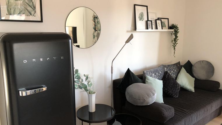 Ein modernes Wohnzimmer mit einer dunklen Couch und gemütlichen Kissen. Im Hintergrund sind ein Kühlschrank und dekorative Bilder an der Wand zu sehen.