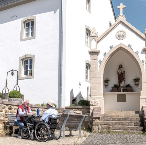 Zwei M&auml;nner, einer im Rollstuhl, sitzen auf einer Bank vor einem wei&szlig;en Geb&auml;ude mit einer religi&ouml;sen Statue in einer Nische. Sonniger Tag., &copy; Naturpark S&uuml;deifel, Thomas Urbany