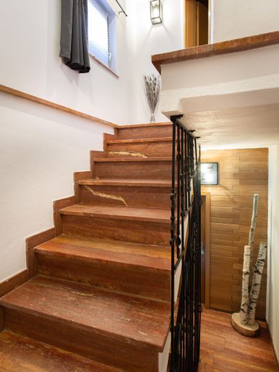 A wooden staircase leads up, surrounded by white walls and a window with curtains. At the end of the staircase is a door, and some decorative elements are visible.
