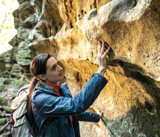 A woman in a blue anorak touches a sandstone wall in the forest. She is carrying a rucksack and looking at the structure of the stone., © Eifel Tourismus GmbH, Dominik Ketz