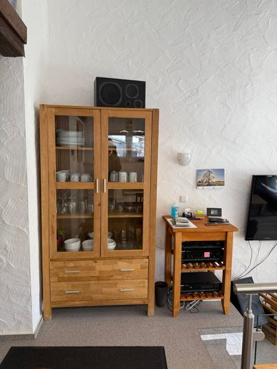 Une armoire en bois avec une façade en verre, qui expose des tasses et de la vaisselle. À côté, il y a une petite table avec un système audio.