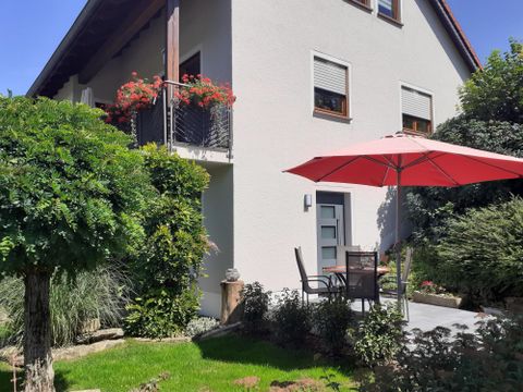 Une belle maison avec un jardin. Une table ronde et des chaises sont sous un parasol rouge.