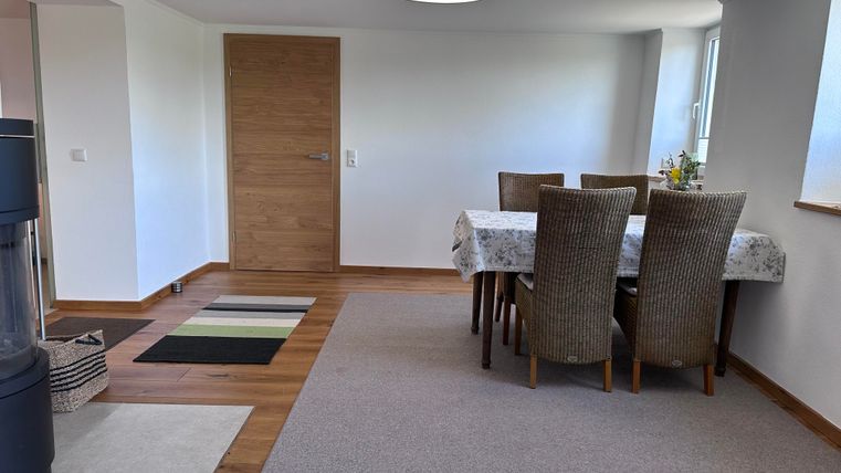 Ein heller, einladender Raum mit einem Tisch und Stühlen. Der Boden ist mit Teppichen ausgelegt und es gibt Fenster, die Tageslicht hereinlassen.