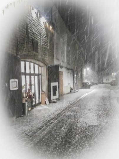 Een met sneeuw bedekte straat met historische gebouwen op de achtergrond. De sneeuw valt zachtjes in de nacht en zorgt voor een romantische sfeer.