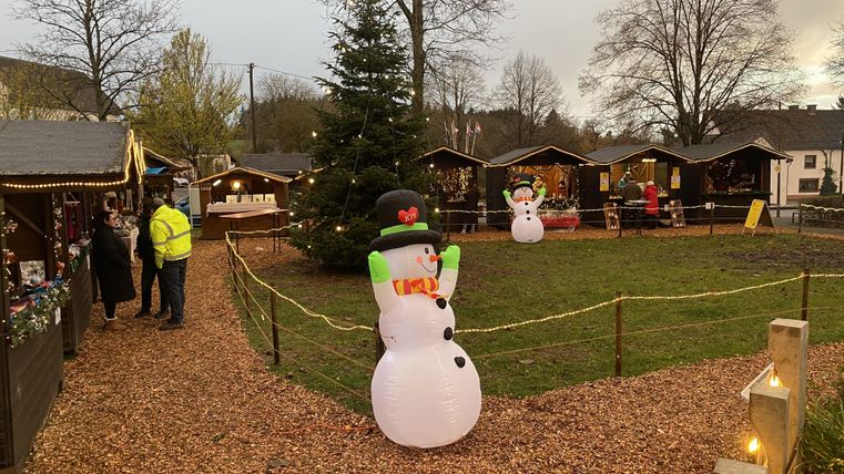 Ein festlich geschmückter Weihnachtsmarkt mit einem großen Weihnachtsbaum und aufblasbaren Schneemännern. Die Umgebung ist von kleinen Hütten und Bäumen umgeben.