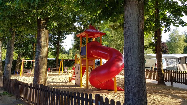 Un aire de jeux avec un toboggan rouge et des balançoires. Entouré d'arbres et d'une clôture en bois.