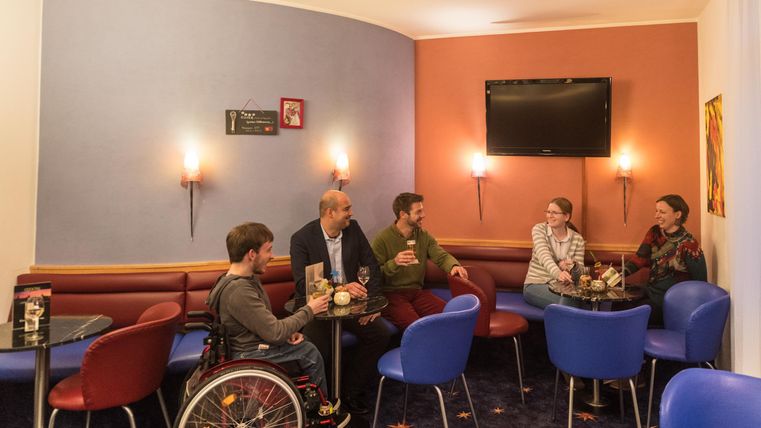 Een groep mensen zit in een gezellige kamer en geniet van drankjes. De zitplaatsen zijn kleurrijk, en op de achtergrond is een televisie te zien.