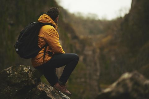 Ein Mann in einer gelben Jacke sitzt auf einem Felsen und schaut in eine Schlucht. Die Landschaft ist grün und mit Bäumen umgeben.