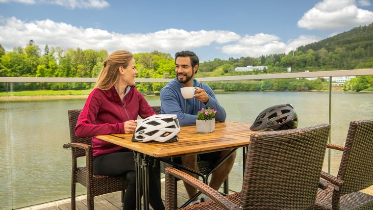 Ein Paar sitzt an einem Tisch am Wasser und genießt Getränke. Im Hintergrund sind grüne Bäume und ein klarer Himmel zu sehen.