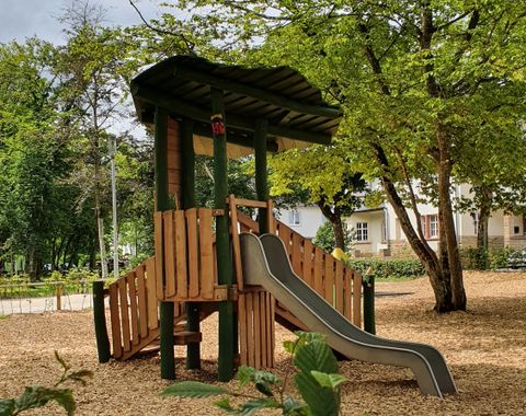 Un aire de jeux avec une structure en bois et un toboggan. La zone est entourée d'arbres et a une ambiance cosy.