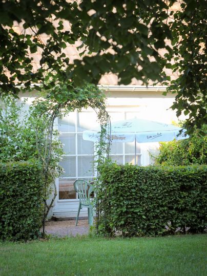 Ein schöner Garten mit einer Sitzgelegenheit unter einem Sonnenschirm. Eine bepflanzte Bogenarchitektur führt zur Eingangstür des Hauses.