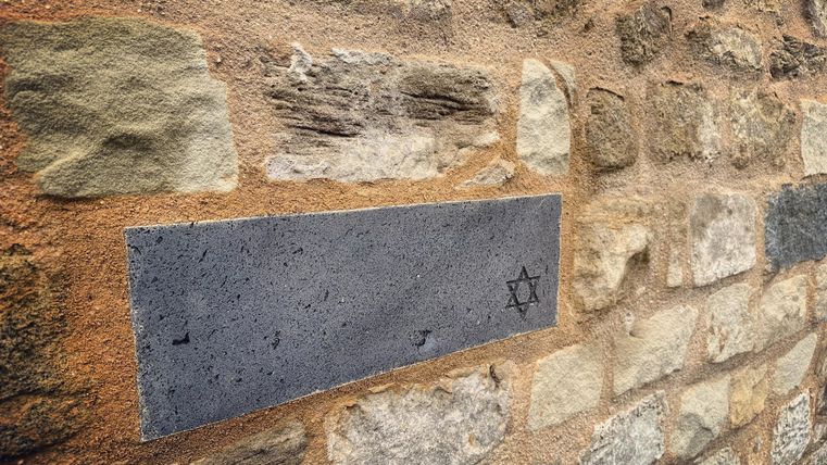 Ein grauer Gedenkstein auf einer Steimmauer mit einem Symbol des Davidsterns. Die Umgebung besteht aus grob bearbeiteten Steinblöcken.