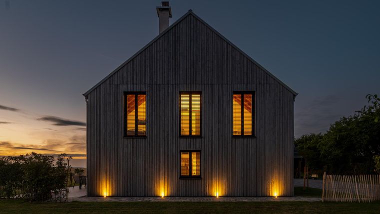 Ein modernes Holzhaus mit beleuchteten Fenstern bei Dämmerung. Im Vordergrund ist ein gut gepflegter Garten zu sehen.