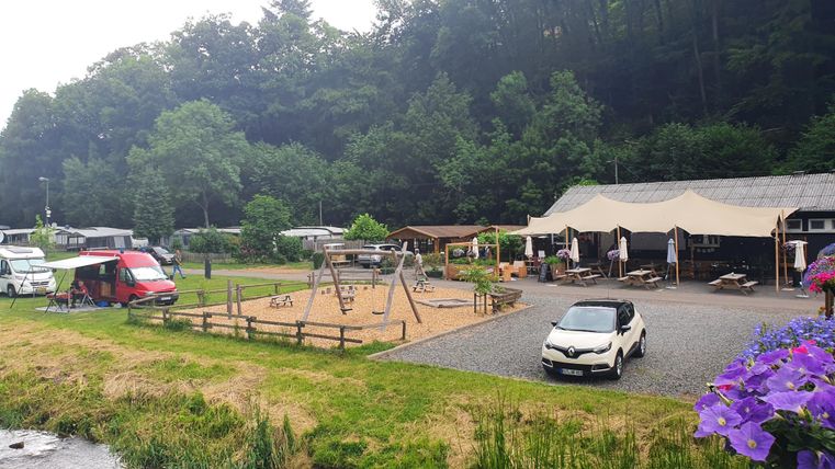 Ein Campingplatz mit Wohnmobilen und einem Spielplatz in einer grünen Umgebung. Im Hintergrund ist ein Gasthaus mit Sitzplätzen im Freien zu sehen.