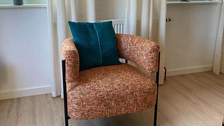 Ein moderner Sessel mit einer bunten Stoffoberfläche und einem blauen Kissen. Im Hintergrund sind Fenster mit Vorhängen und eine kleine Pflanze zu sehen.