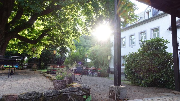 Une cour accueillante avec des sièges et beaucoup de verdure. Le soleil brille à travers les arbres et crée une atmosphère chaleureuse.