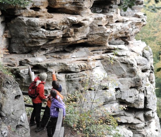 Three hikers on a path in the Devil's Gorge, looking at and pointing to impressive rock formations., © Felsenland Südeifel Tourismus GmbH