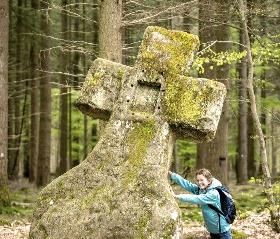 Girl in blue jacket leaning against the moss-covered Fraubille cross in the forest of Bollendorf., © Eifel Tourismus GmbH, Dominik Ketz