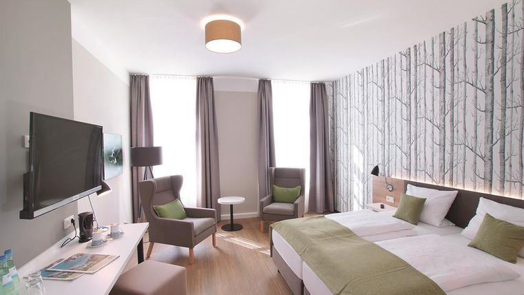 A modern hotel room with a double bed and elegant furnishings. There are windows with curtains, a flat-screen TV, and a cozy seating area.