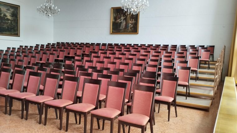 Ein großer Raum mit vielen Reihen von pinken Stühlen. Im Hintergrund sind Kunstwerke und Kristallleuchter zu sehen.