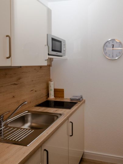 Eine moderne Küche mit einer Edelstahlspüle, einem Induktionskochfeld und einer Mikrowelle. Die Wände sind hell und das Holzdekor verleiht eine warme Atmosphäre.