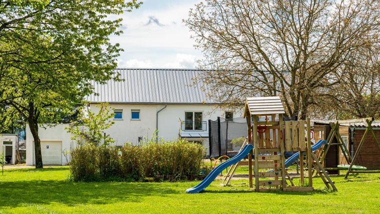 Ein Spielplatz mit Rutsche und Spielgerüst in einer grünen Wiese. Im Hintergrund ist ein helles Gebäude und einige Bäume zu sehen.