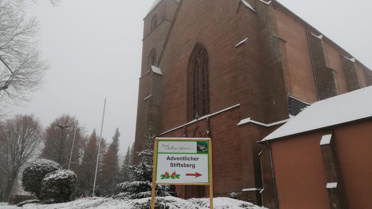 Eine Kirche im Schnee mit einem Schild, das auf den Adventlichen Stiftsberg hinweist. Die Landschaft ist winterlich und lädt zum Besuch ein.