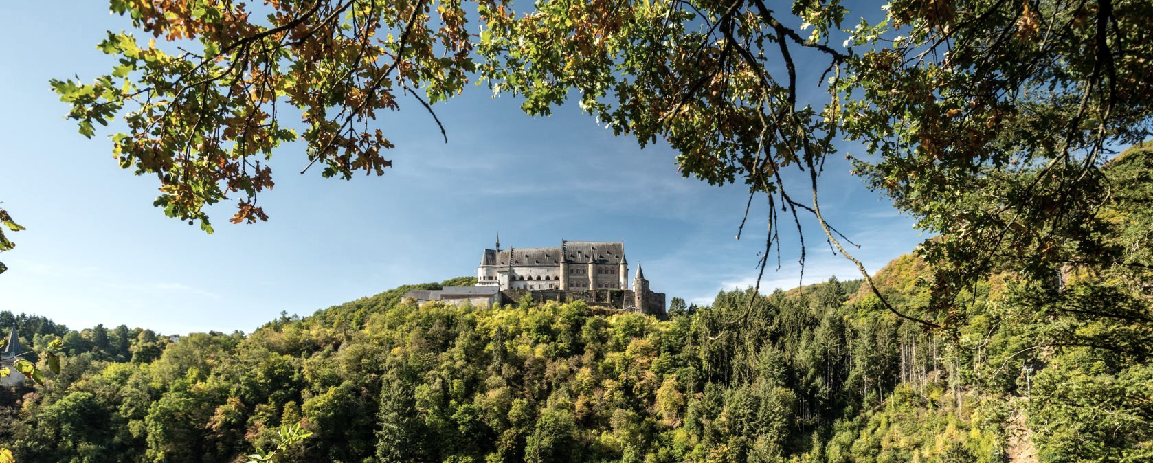 Schloss Vianden thront majestätisch auf einem bewaldeten Hügel, umgeben von herbstlichen Bäumen und blauem Himmel., © Eifel Tourismus GmbH, D. Ketz
