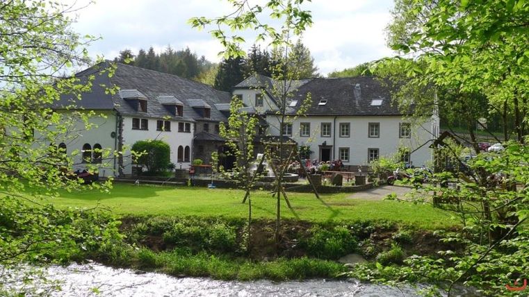 Ein charmantes Gebäude in einer grünen Umgebung mit einem Fluss im Vordergrund. Die Terrasse ist mit Blumen und Sitzgelegenheiten ausgestattet.
