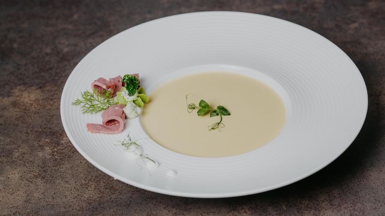 Une soupe crémeuse dans une assiette blanche, élégamment présentée. À côté de la soupe se trouvent du jambon et des herbes fraîches.