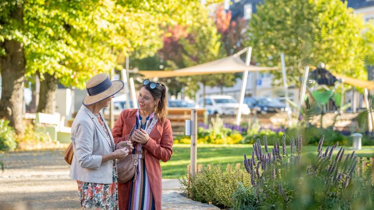 Zwei Frauen unterhalten sich in einem schönen Park mit grünen Bäumen und bunten Blumen. Die Sonne scheint und die Atmosphäre ist freundlich und entspannt.