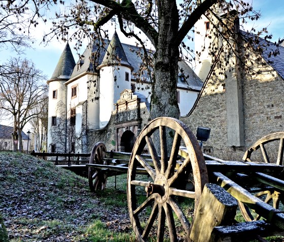 Rittersdorf Castle in winter, surrounded by bare trees. In the foreground, old wooden wagon wheels create a historic atmosphere., © TI Bitburger Land