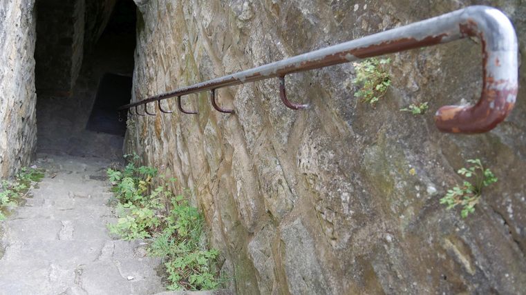 Eine steinerne Treppe führt hinunter zu einem dunklen Durchgang. An der Wand gibt es ein rostiges Geländer.