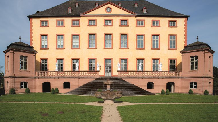 Ein beeindruckendes Schloss mit einer orangefarbenen Fassade und großen Fenstern. Es liegt umgeben von einem gepflegten Garten und bietet eine majestätische Aussicht.