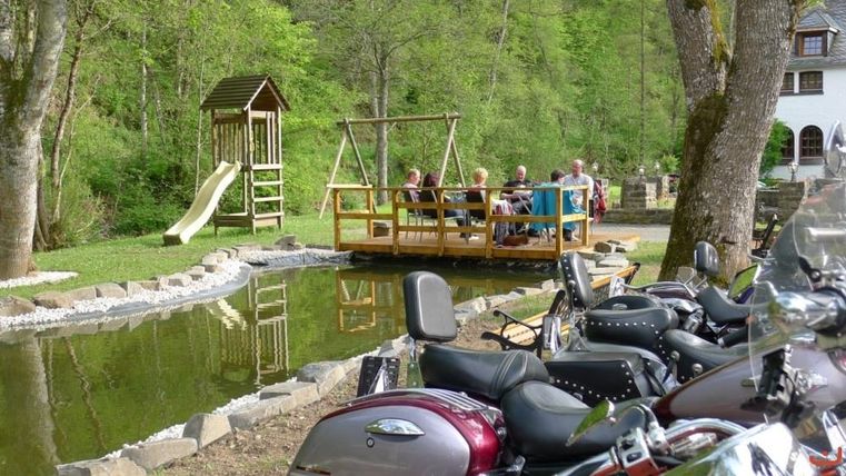A relaxed garden landscape with a playground and a terrace by the water. In the foreground, motorcycles are parked.