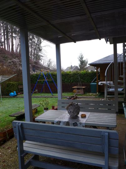 Ein gemütlicher Garten mit einem Holztisch und Bänken. Im Hintergrund sind ein Spielplatz und ein Pavillon sichtbar.