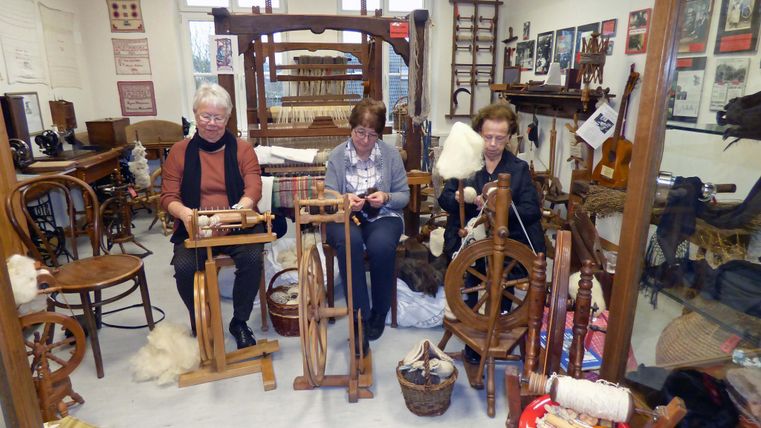 Drei Frauen spinnen Wolle an Spinnrädern in einem traditionellen Handwerksraum. Im Hintergrund sind verschiedene Werkzeuge und historische Bilder zu sehen.