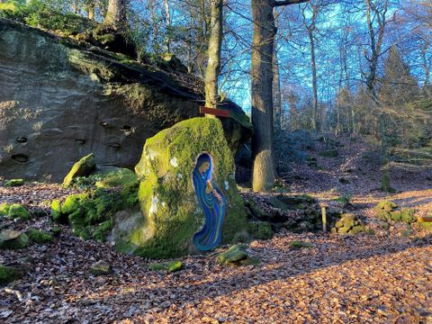 Ein bunter Fels mit einer künstlerischen Darstellung in einem Wald. Umgeben von Laub und hohen Bäumen im Sonnenlicht.