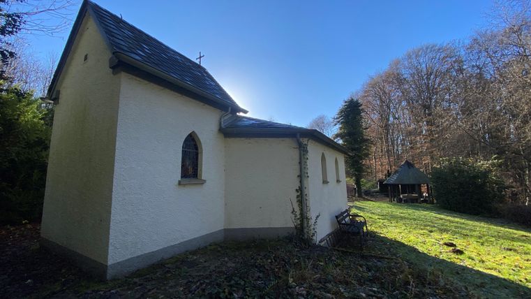 Eine kleine Kirche mit einem hellen Fassadenanstrich. Im Hintergrund sind Bäume und eine Wiese zu sehen.