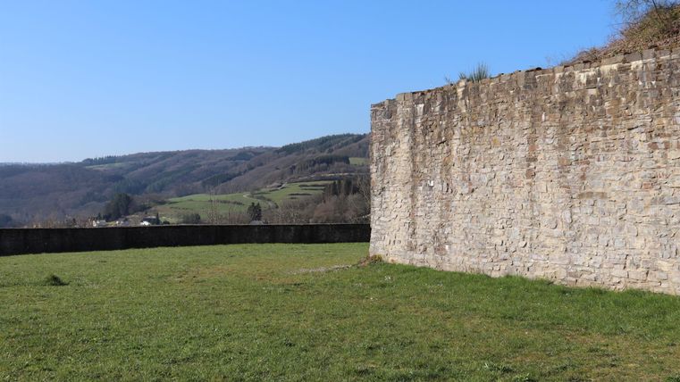 Eine alte Steinmauer steht in einem grünen Feld unter klarem blauen Himmel. Im Hintergrund sind sanfte Hügel zu sehen.
