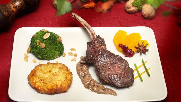 Une assiette joliment dressée avec des côtelettes d'agneau, du brocoli et un gratin de pommes de terre. Des accompagnements fruités et une garniture décorative complètent le plat.