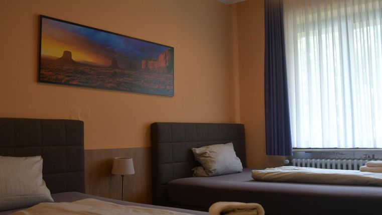 Een eenvoudige kamer met twee bedden en een schilderij aan de muur. Er zijn grote ramen die veel daglicht binnenlaten.