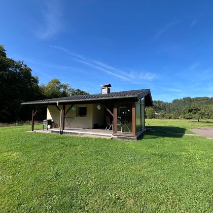 Ein kleines, gemütliches Haus steht in einer grünen Wiese. Der Himmel ist blau und klar, umgeben von Bäumen.