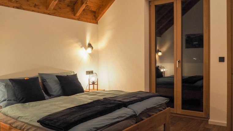 Ein modernes Schlafzimmer mit einem Holzstuhl und einem großen Bett. Die Wände sind in neutralen Tönen gehalten, und große Fenster reflektieren das Licht.