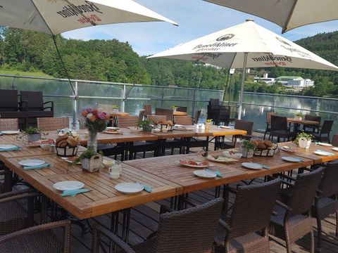 An inviting outdoor dining area with several table settings. The view overlooks a serene lake and green forests.